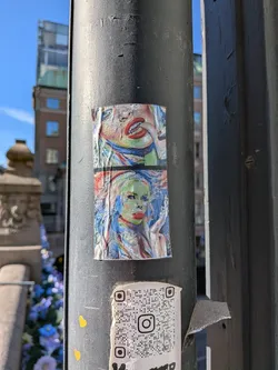 Street sticker The central sticker is a two-part artwork, vertically stacked, depicting abstract and expressionistic portraits. The upper section features a close-up of a mouth with vivid red lips, slightly open, revealing teeth, set against a textured background with a blend of blue, green, and yellow tones. The lower section portrays a stylized face, likely female, with striking red lips, prominent eyes, and a complexion rendered in shades of green and yellow, framed by flowing blue hair. The artwork exhibits a painterly style with visible brushstrokes, creating a vibrant and somewhat distressed aesthetic. The sticker itself is creased, wrinkled, and shows signs of wear, indicating its physical presence on the metal pole.