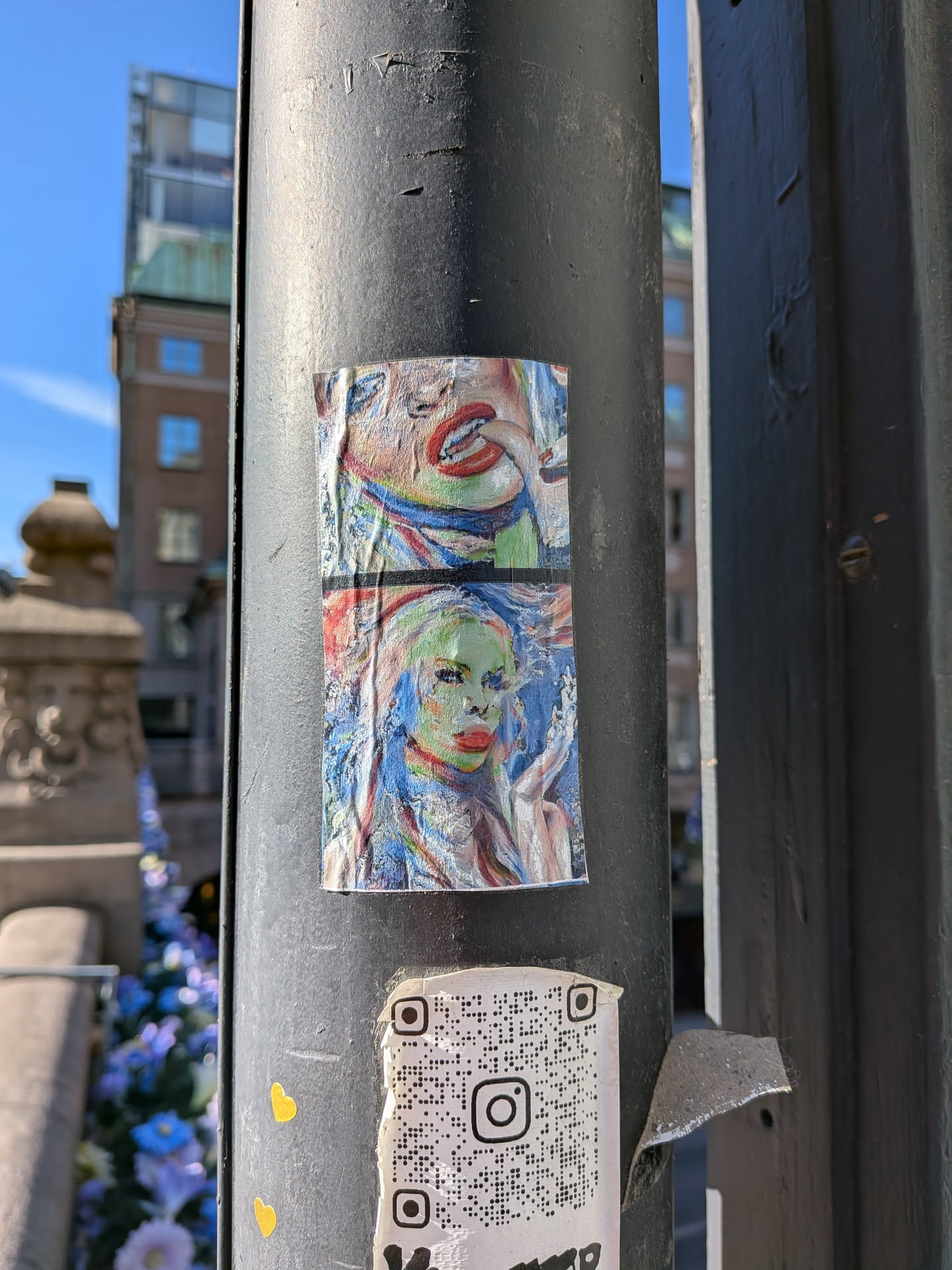 The central sticker is a two-part artwork, vertically stacked, depicting abstract and expressionistic portraits. The upper section features a close-up of a mouth with vivid red lips, slightly open, revealing teeth, set against a textured background with a blend of blue, green, and yellow tones. The lower section portrays a stylized face, likely female, with striking red lips, prominent eyes, and a complexion rendered in shades of green and yellow, framed by flowing blue hair. The artwork exhibits a painterly style with visible brushstrokes, creating a vibrant and somewhat distressed aesthetic. The sticker itself is creased, wrinkled, and shows signs of wear, indicating its physical presence on the metal pole.