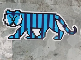 A blue and dark blue pixel art tiger sticker. The tiger is depicted in profile, walking to the right. It has a striped pattern and is somewhat cartoonish in style. The sticker is affixed to a textured gray surface.