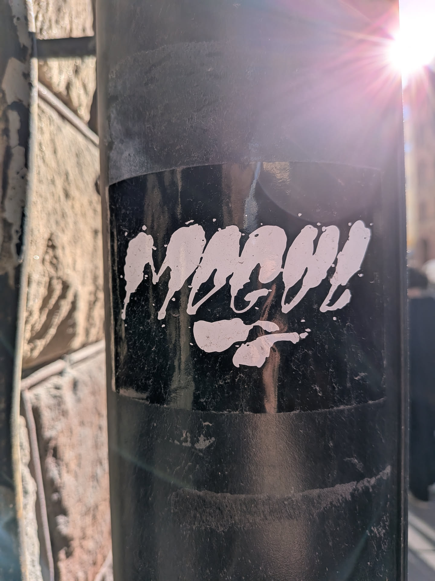The sticker is a rectangular piece with a matte black background, applied to a dark, possibly metal, pole. The central design features highly stylized, white, paint-splatter-like typography that appears to spell 'Mmm!!'. The letters have an organic, drippy, and distressed texture, with numerous small white flecks scattered around them, mimicking a stencil or spray paint effect. The sticker shows signs of wear, with slight scuffing and a somewhat faded appearance, particularly visible under the bright sunlight reflecting from the top right.