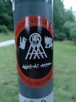 Street sticker Black and orange round sticker with the text "Allt ät Alla" in the center, surrounded by the phrase "För och en efter för" in a circular pattern. The central design includes a stylized image with what looks like a rollercoaster or similar structure. The sticker is attached to a gray pole.