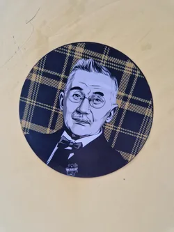 Street sticker This circular sticker showcases a detailed illustration of a man, possibly a historical figure or a fictional character, with glasses and a bow tie.  He's depicted in a grayscale style, creating a contrast against the dark and gold plaid pattern. The sticker seems to be high-quality and well-designed.