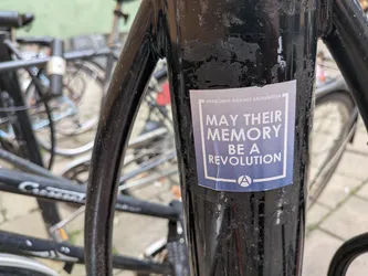 Street sticker A rectangular sticker with a blue-grey background and white text, affixed to a black metal pole. The main text reads 'MAY THEIR MEMORY BE A REVOLUTION' in a bold, white, sans-serif font, framed by a white border. Above the border, smaller text reads 'ANARCHISTS AGAINST ANTISEMITISM'. At the bottom, a white circle contains the letter 'A', representing the anarchy symbol. The sticker is slightly worn and peeling at the bottom-left corner.