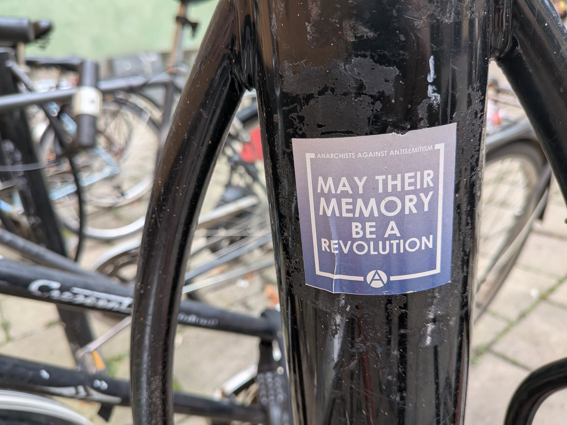 A rectangular sticker with a blue-grey background and white text, affixed to a black metal pole. The main text reads 'MAY THEIR MEMORY BE A REVOLUTION' in a bold, white, sans-serif font, framed by a white border. Above the border, smaller text reads 'ANARCHISTS AGAINST ANTISEMITISM'. At the bottom, a white circle contains the letter 'A', representing the anarchy symbol. The sticker is slightly worn and peeling at the bottom-left corner.