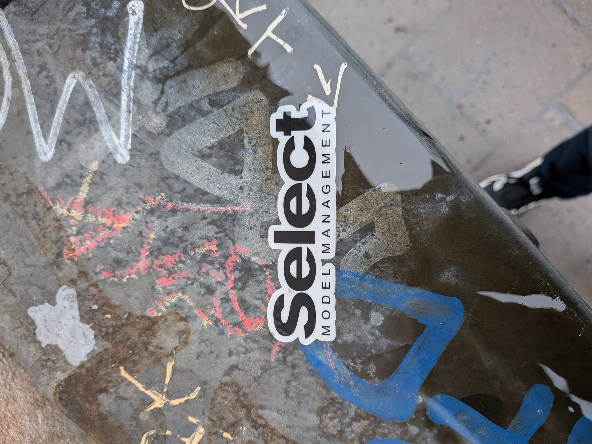 The sticker is a rectangular shape with slightly rounded corners, featuring a clean, minimalist design. It has a white background with black text. The prominent word "Select" is written vertically in a bold, sans-serif typeface, with the 'S' at the bottom and 't' at the top. To the right of "Select", the words "MODEL MANAGEMENT" are stacked vertically in a smaller, thinner, all-caps sans-serif font. The sticker appears to be slightly worn around the edges, indicating it's a physical adhesive label.