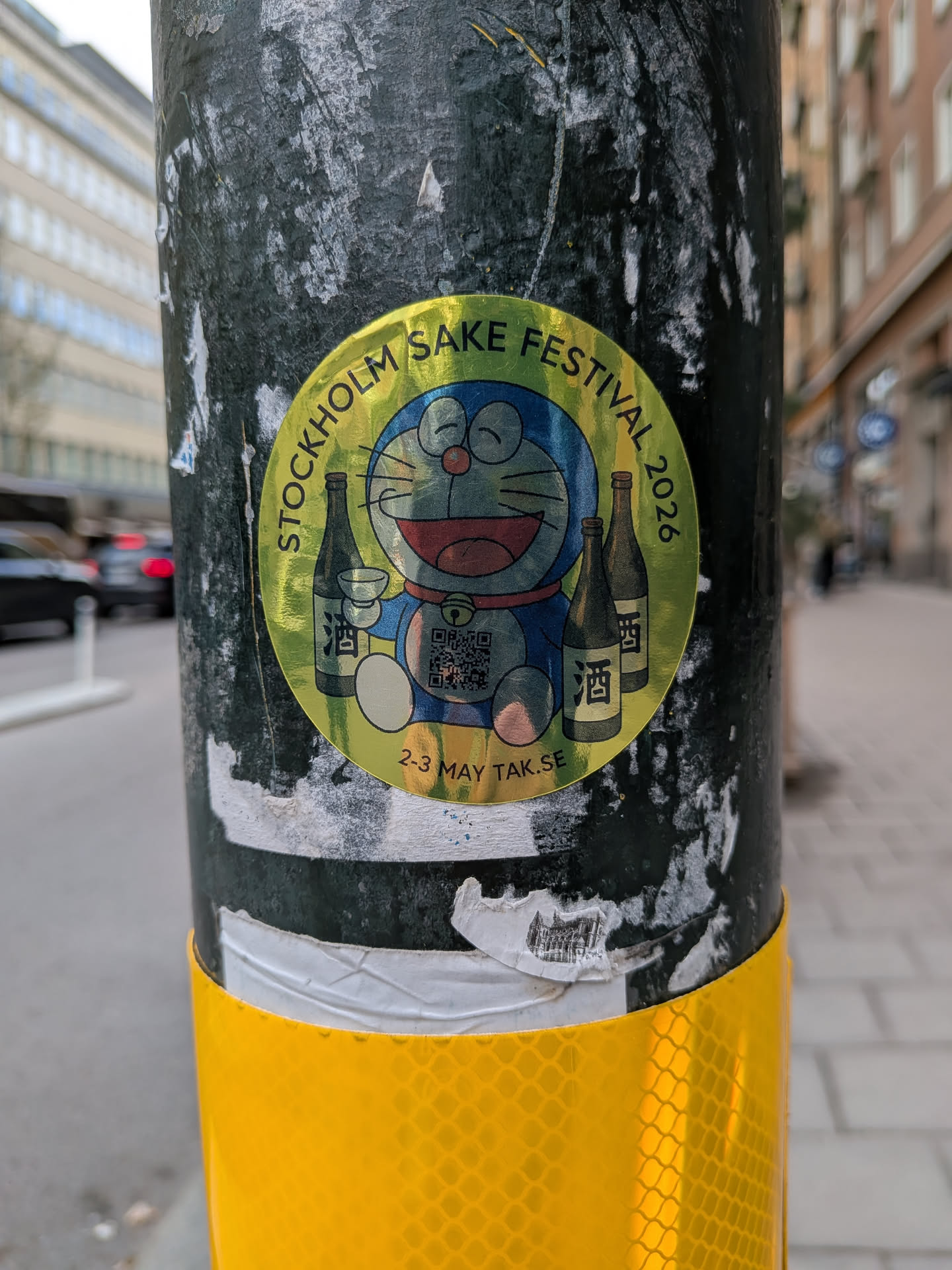 A circular, metallic golden-yellow sticker featuring the popular blue and white anime character Doraemon. Doraemon is depicted with a wide smile, holding a small sake cup. Flanking him are two traditional brown sake bottles, each adorned with the Japanese character for 'sake' (酒). The top arc of the sticker reads 'STOCKHOLM SAKE FESTIVAL 2026' in black capital letters. At the bottom, '2-3 MAY TAK.SE' is written. A QR code is prominently displayed on Doraemon's belly area. The sticker has a shiny, slightly holographic appearance.