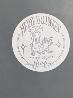 Street sticker This is a round sticker featuring cartoon beer characters. The sticker has the text "Heide Halunken" written in an arc at the top. Below the text, there are two cartoon characters: one resembling a glass of beer with a frothy head and the other a smaller glass with a red liquid inside, both with arms and legs. Below the characters, it reads "MATCH MADE AT Heide". There are star shapes on both sides of the cartoon characters.