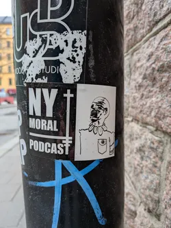 Street sticker The sticker is a rectangular white sticker featuring a black line drawing of a character from the chest up. The character has a somewhat grotesque or unsettling appearance, with a wide, open mouth revealing teeth, and large, dark eyes. Its head is bald or has no discernible hair. Around its neck, it wears a distinctive collar made of what appears to be rounded, possibly bone-like or tribal elements, connected by a string. The character is depicted wearing a shirt with a single breast pocket on the left side, which has a small, simple flower-like design drawn within it. The drawing style is simple, bold line art, giving it a somewhat cartoonish yet eerie quality. The sticker is predominantly white with black lines for the illustration.