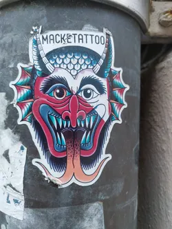 A vibrant sticker featuring a traditional-style tattoo of a devil's head. The artwork is detailed and colorful, with sharp lines and bold colors. The devil's expression is fierce, yet playful. The sticker is affixed to a dark gray surface, adding contrast to the design.