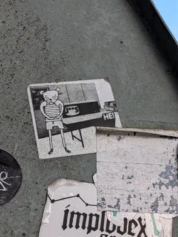 Street sticker The sticker features a black and white illustration of a cartoon bear standing in front of a table. The bear has large eyes, a small snout, and is wearing a horizontally striped shirt and shorts. Its arms are crossed over its chest, and its legs are thin with simple feet. Behind the bear, a table is depicted with a checkered pattern on its surface. To the right of the bear, the text 'HE!' is visible. The sticker appears to be slightly worn and has a textured, pixelated background. It's positioned on a rough, weathered surface, contributing to its overall distressed aesthetic.