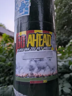 Street sticker The sticker is rectangular with slightly rounded corners, adhered to a dark green pole. The top portion features a banner with red and yellow stripes, reminiscent of a flag. Prominently displayed are the words "GO AHEAD" in a bold, stylized font; "GO" is red, and "AHEAD" is yellow, both outlined in black and white. To the right of "AHEAD" is a grey, stylized trophy cup. Below "GO AHEAD" in smaller white text is the Dutch phrase "IK HOU VAN JOU HET MEEST" (I love you the most). The central and largest part of the sticker depicts a vibrant football stadium scene, likely during a night match. The stands are visible with spectators, and the field below is engulfed in thick white smoke from numerous red flares or pyrotechnics lit along the lower stands. Football players are visible on the pitch. Small, partially obscured logos like "TOTO", "Heineken", and "KNVB BEKER" can be seen within the stadium image. The bottom of the sticker has a black band with red text that reads "BEKERWINNAAR 2025" (Cup Winner 2025), indicating a future aspiration.