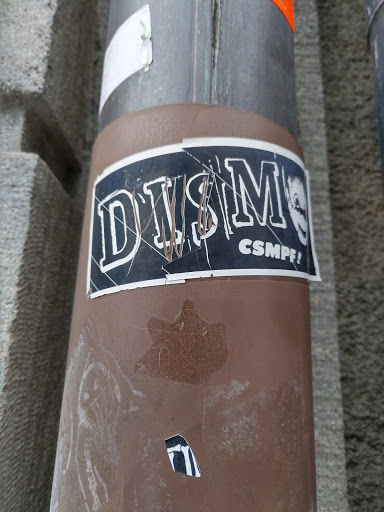 A sticker with the words "DYSMORPHIA CSMPF!" on a black background.  The sticker is affixed to a brown pole and is somewhat damaged.