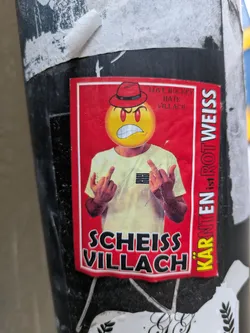 Street sticker Sticker featuring an angry emoticon wearing a red hat and giving the middle finger. The text on the sticker says "Love hockey hate Villach" and "Kärnten ist not Weiss" in German, which translates to "Carinthia is not white". There is also the phrase "Scheiss Villach", which means "Shit Villach".
