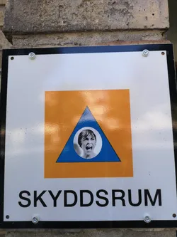 Street sticker A circular black-and-white sticker of Janet Leigh screaming from the movie 'Psycho', pasted in the center of a blue triangle on a Swedish 'SKYDDRUM' shelter sign.
