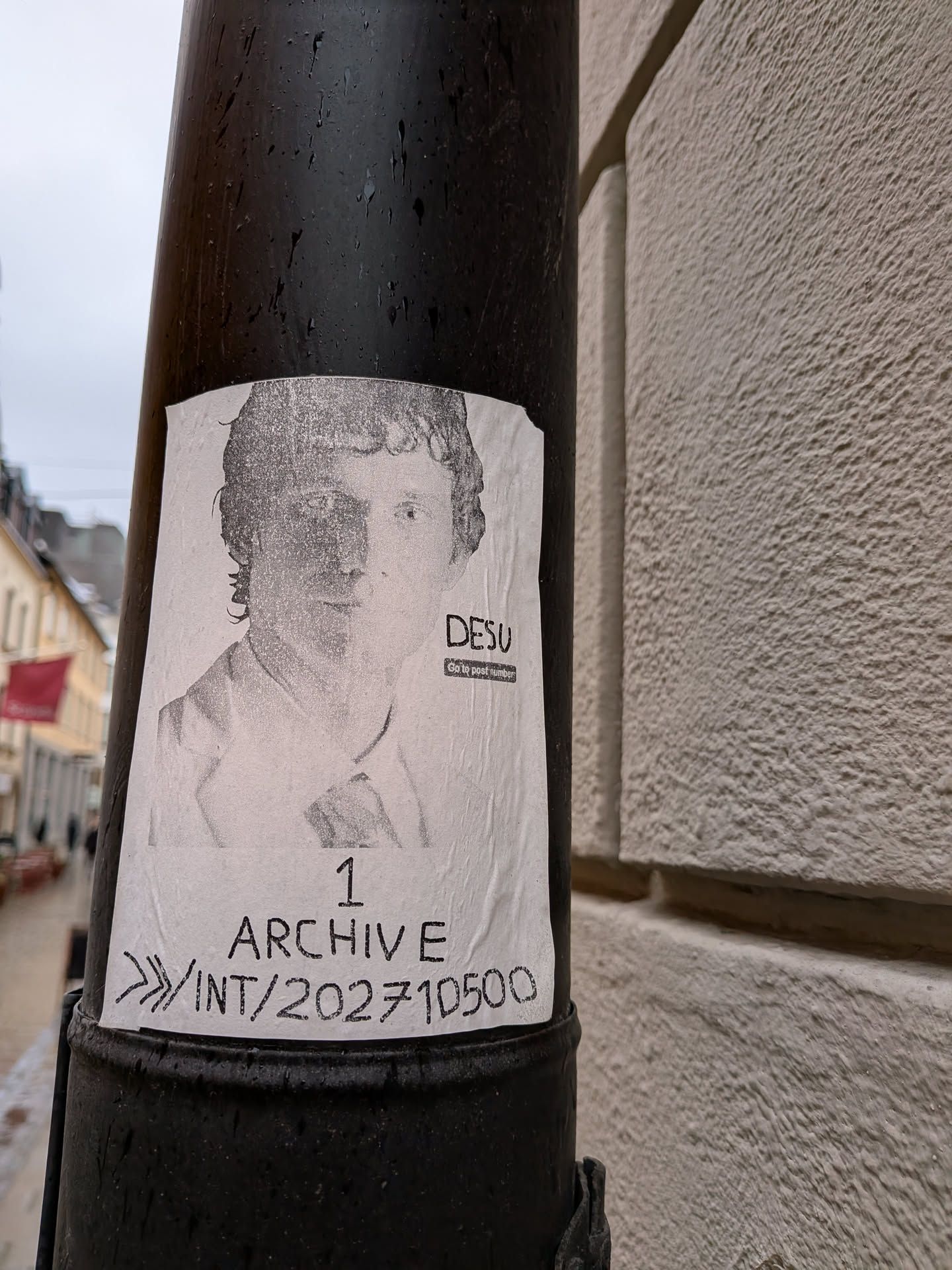 The sticker is a rectangular, white paper print affixed to a dark, textured pole. It features a grainy, black-and-white portrait of a man with curly hair, looking forward. His left side is partially obscured by shadow. He appears to be wearing a collared shirt and a jacket. Below the portrait, there is text in a handwritten-style font. The paper shows signs of wear, including wrinkles and a slightly curled bottom edge, indicating it's a real-world paste-up or sticker.