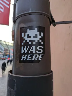 The sticker is black with white pixelated graphics. At the top, it features a classic Space Invader character. Below the character, the text 'WAS HERE' is displayed in a pixelated font, mirroring the style of the invader. The sticker appears to be applied to a dark, cylindrical object, possibly a lamppost or utility pole.