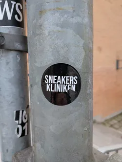 Street sticker A circular black sticker with bold white uppercase text. The word 'SNEAKERS' is on the top line, and 'KLINIKEN' is on the bottom line. The sticker has a slightly glossy finish, showing minor reflections, and is adhered to a textured, weathered grey metal pole.