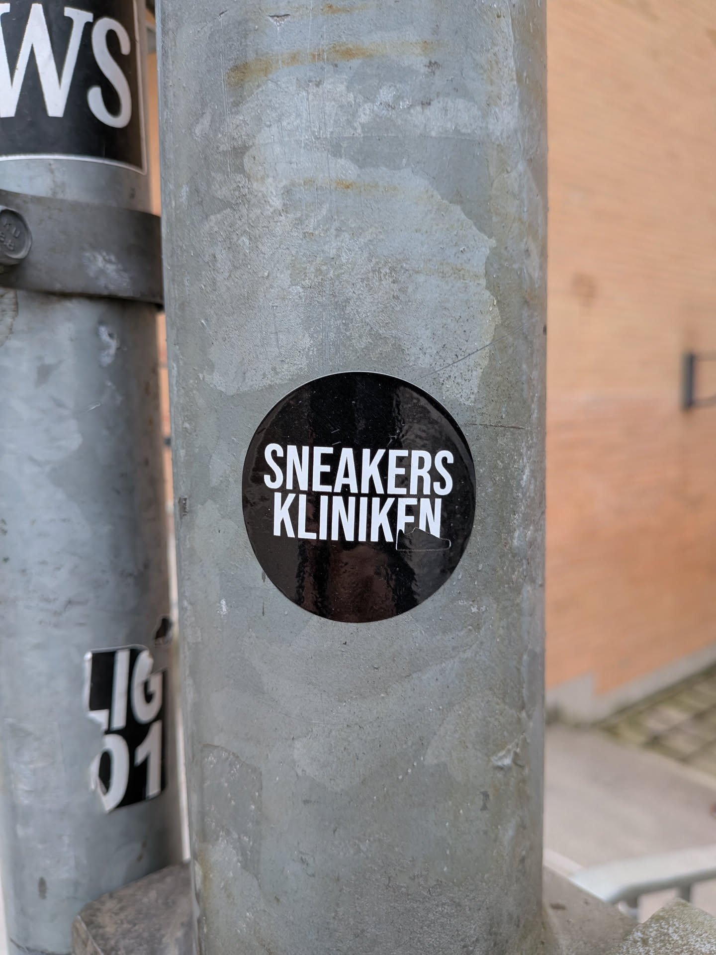 A circular black sticker with bold white uppercase text. The word 'SNEAKERS' is on the top line, and 'KLINIKEN' is on the bottom line. The sticker has a slightly glossy finish, showing minor reflections, and is adhered to a textured, weathered grey metal pole.