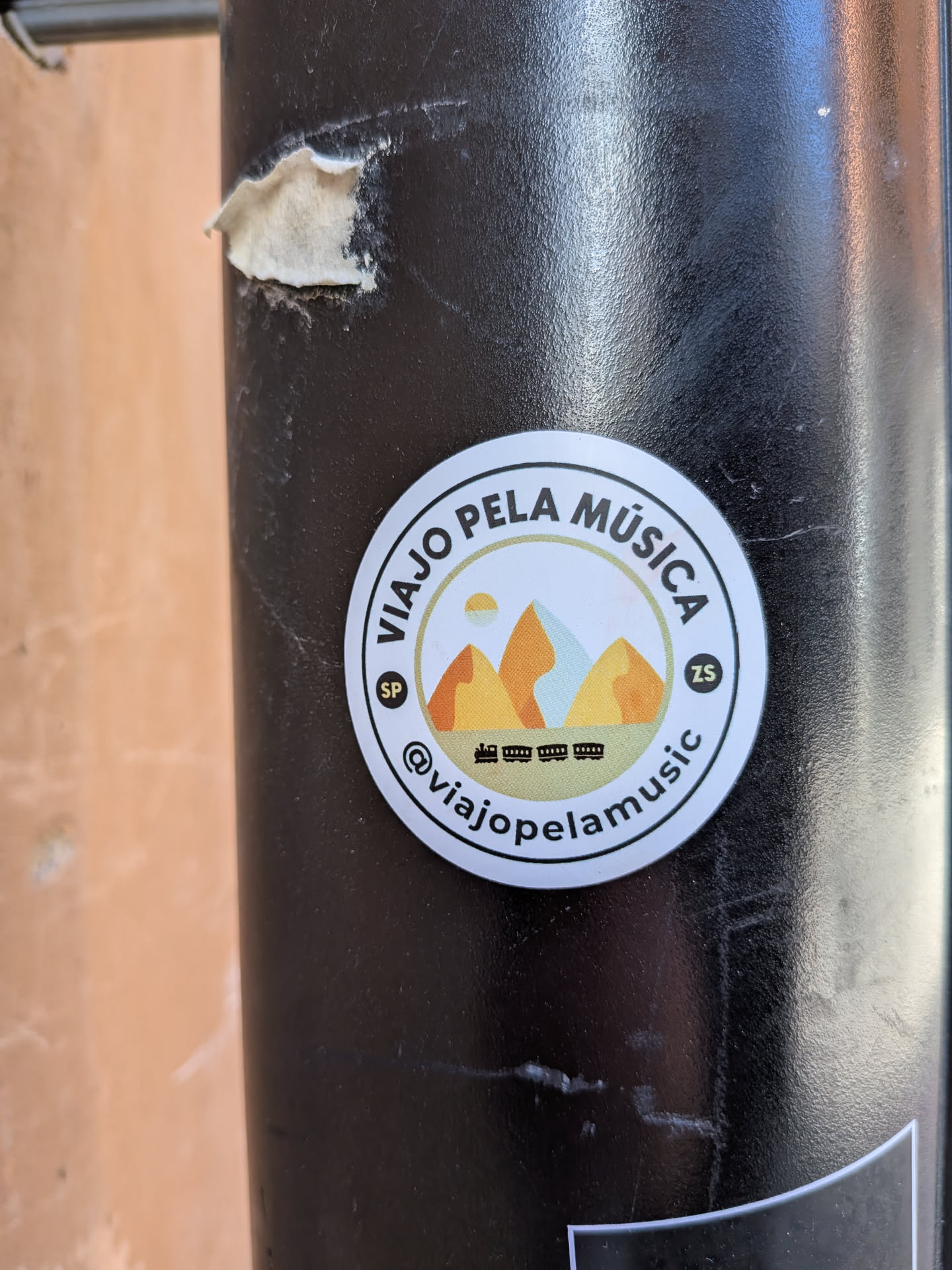 A circular sticker featuring a stylized landscape design. The central image depicts three prominent mountains in shades of orange and light blue, with a small yellow sun or moon partially visible behind the leftmost mountain. Below the mountains, a green strip represents the ground, upon which a black silhouette of a train with multiple carriages is visible. The design is framed by a thin yellow-green circle, then a black line, and finally a white outer border. Arched text at the top reads "VIAJO PELA MÚSICA" in black, and at the bottom, "@viajopelamusic" is also in black. Small black circles with white text "SP" and "ZS" are positioned on the left and right sides of the central image, respectively.