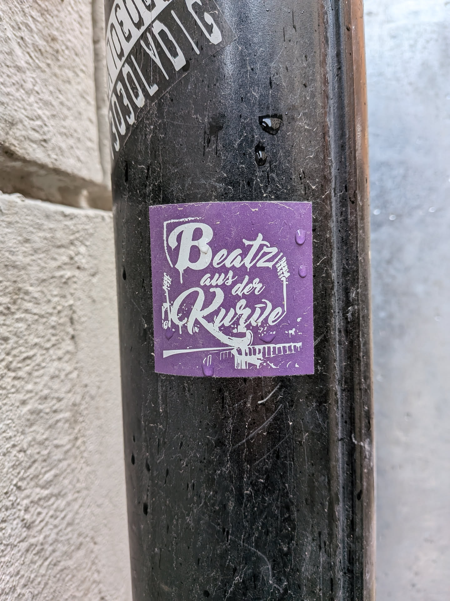 A square-shaped sticker with a vibrant purple background and a white design. The central text reads 'Beatz aus der Ruhr' in a stylized, somewhat drippy script font. 'Beatz' and 'Ruhr' are prominently displayed, with 'aus der' in smaller text between them. To the left of the text is a stylized illustration of an electric guitar, and to the right, a microphone stand. Below the text, a silhouette of an industrial landscape or bridge structure is depicted. The overall aesthetic is reminiscent of street art or music event branding, with water droplets visible on its surface, indicating it's outdoors.