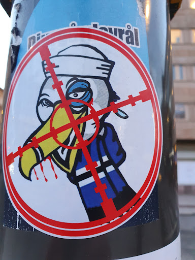 The sticker depicts a cartoon toucan in a sailor's hat, seemingly targeted by a crosshair. It has a dark and slightly morbid aesthetic.  The background is partially visible and seems to show another sticker.