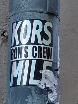 A holographic sticker featuring the text "KORS RON'S CREW MILE" on a black background. The sticker is attached to a grey metal pole.