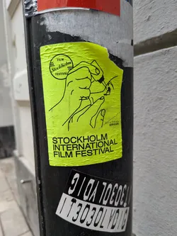 Street sticker The sticker is a bright, almost neon, yellow-green rectangle with black line art and text. It features a stylized illustration of two hands. The upper hand appears to be holding or interacting with a circular object, possibly a film reel, while the lower hand is positioned beneath it. In the upper left corner, there's a circular logo containing the words 'FILM Stockholm FESTIVAL'. To the right of the main illustration, there is Japanese text. Below the illustration, the words 'STOCKHOLM INTERNATIONAL FILM FESTIVAL' are prominently displayed in black capital letters. The sticker shows signs of wear, including wrinkles and slight tears, indicating it's a physical sticker adhered to a surface.