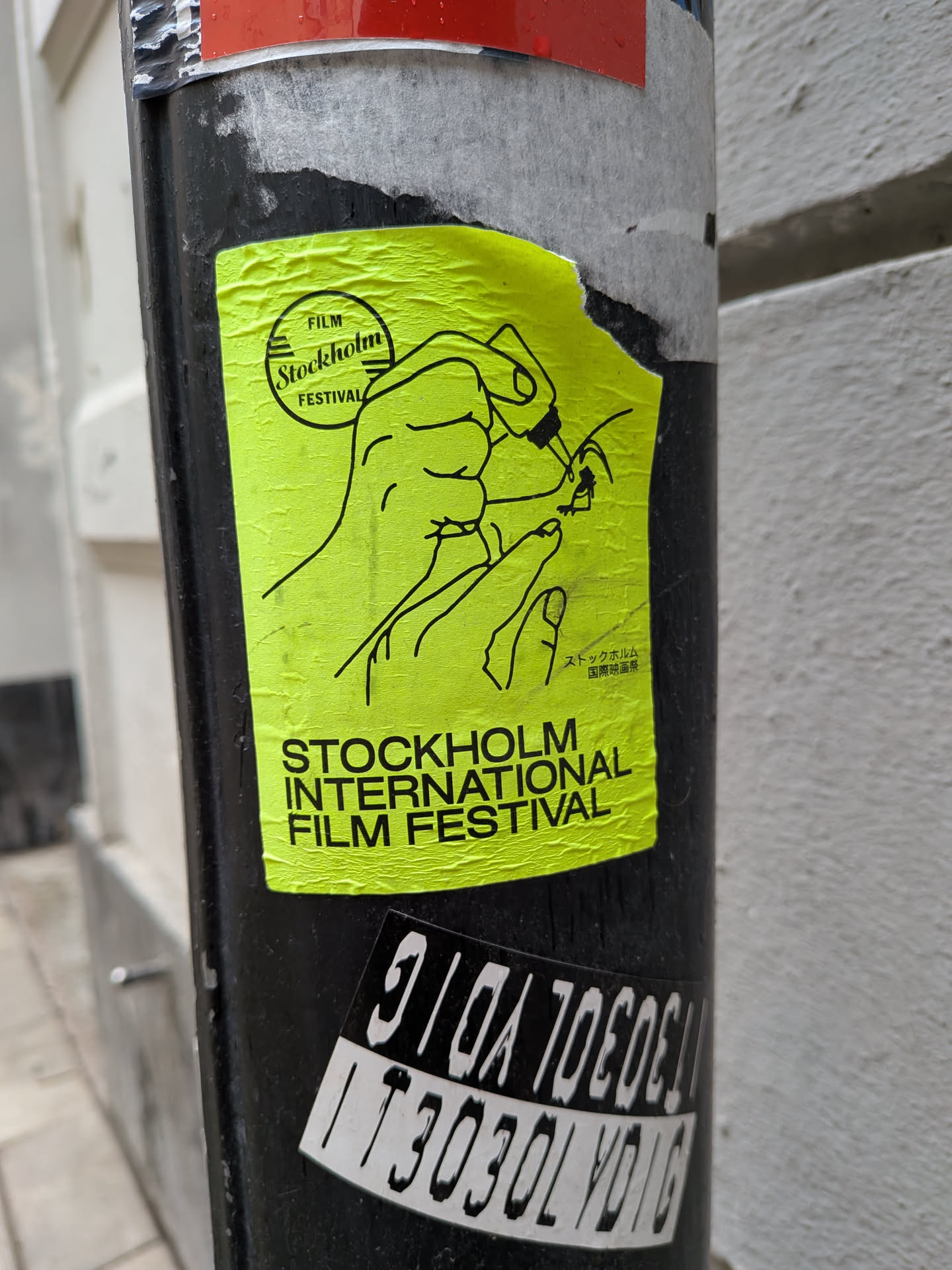 The sticker is a bright, almost neon, yellow-green rectangle with black line art and text. It features a stylized illustration of two hands. The upper hand appears to be holding or interacting with a circular object, possibly a film reel, while the lower hand is positioned beneath it. In the upper left corner, there's a circular logo containing the words 'FILM Stockholm FESTIVAL'. To the right of the main illustration, there is Japanese text. Below the illustration, the words 'STOCKHOLM INTERNATIONAL FILM FESTIVAL' are prominently displayed in black capital letters. The sticker shows signs of wear, including wrinkles and slight tears, indicating it's a physical sticker adhered to a surface.