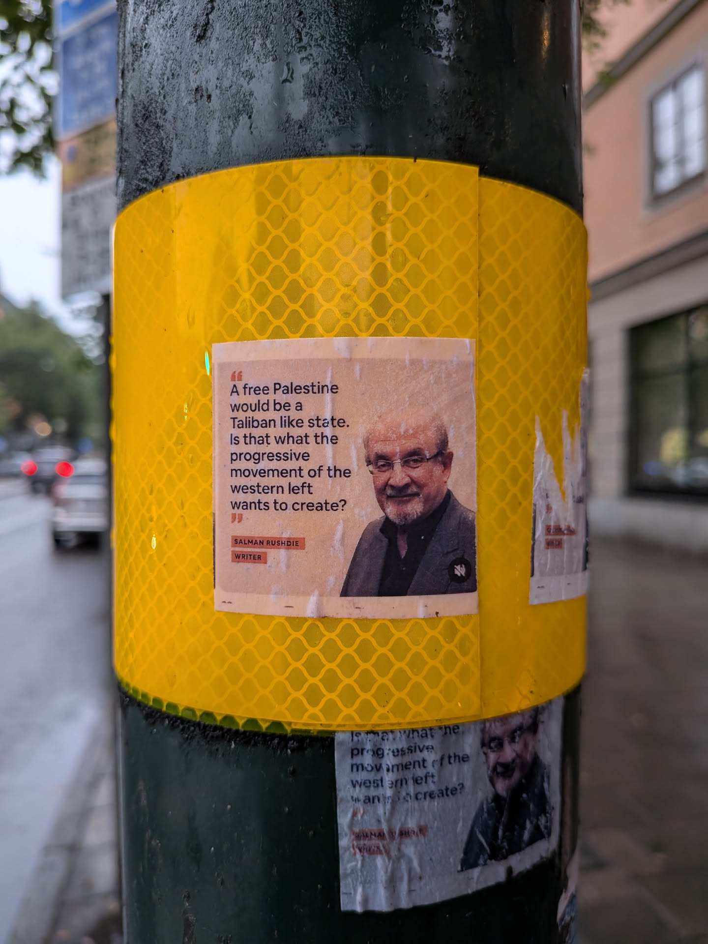 Square paper sticker on a yellow pole. Features a portrait of Salman Rushdie alongside a political quote questioning the Western left's stance on Palestine.