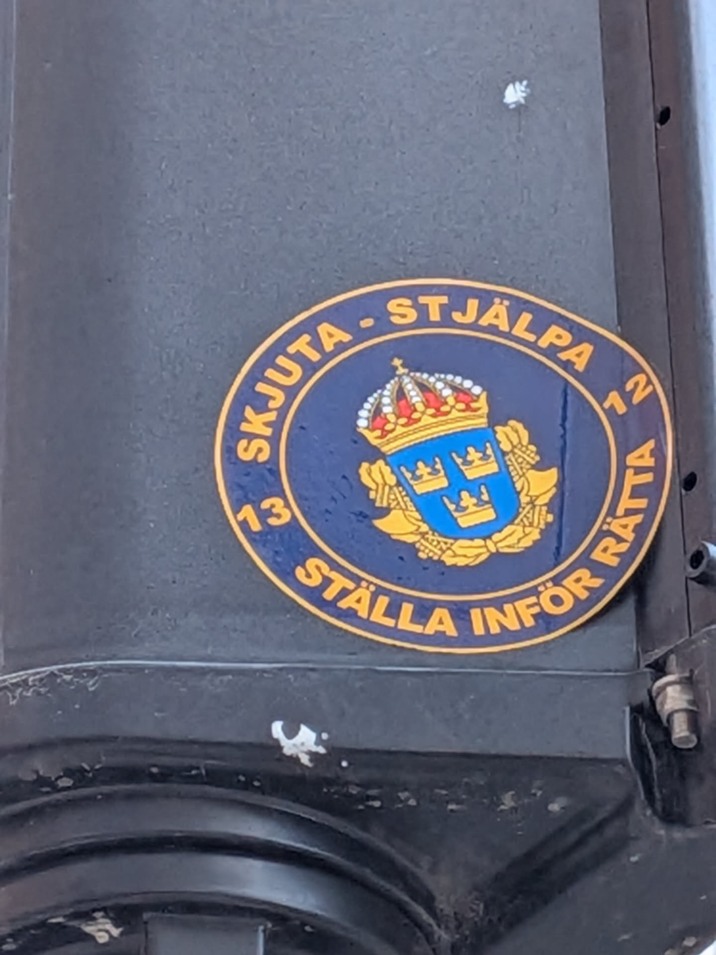 Street sticker A circular sticker with a dark blue background and a gold/yellow border. At its center is the Lesser Coat of Arms of Sweden, depicted as a blue shield bearing three gold crowns (two above one). Above the shield rests a large, ornate royal crown with red and white jewel details. Gold/yellow laurel-like branches or ornamentation frame the shield. Along the outer gold border, text is written in gold/yellow capital letters. The top arc reads "SKJUTA - STJÄLPA", the right side has the number "12", the bottom arc reads "STÄLLA INFÖR RÄTTA", and the left side has the number "13". The sticker is adhered to a dark, textured surface, possibly a metal pole or casing.