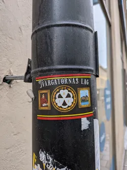 Street sticker The sticker is rectangular with a black background, bordered by a red stripe at the top and a red and yellow stripe at the bottom. The top features the text 'Tvärgatornas Lag' in white capital letters. Below this, the sticker is divided into three main visual elements. On the left, a framed portrait depicts a man with glasses, wearing a brown jacket, seated at a desk with papers, identified by the name 'Nils Norin' below. In the center, a prominent gold-bordered circular emblem showcases a white three-leaf clover or shamrock, with the letters 'B', 'I', and 'F' inscribed within its leaves, and the year '1912' above. On the right, another framed image displays a painting or illustration of a large, multi-story house or villa situated by water, with a flag flying from its roof, identified as 'Villa Rettig' below. The overall aesthetic is classic and commemorative, utilizing gold and white accents against the dark background.