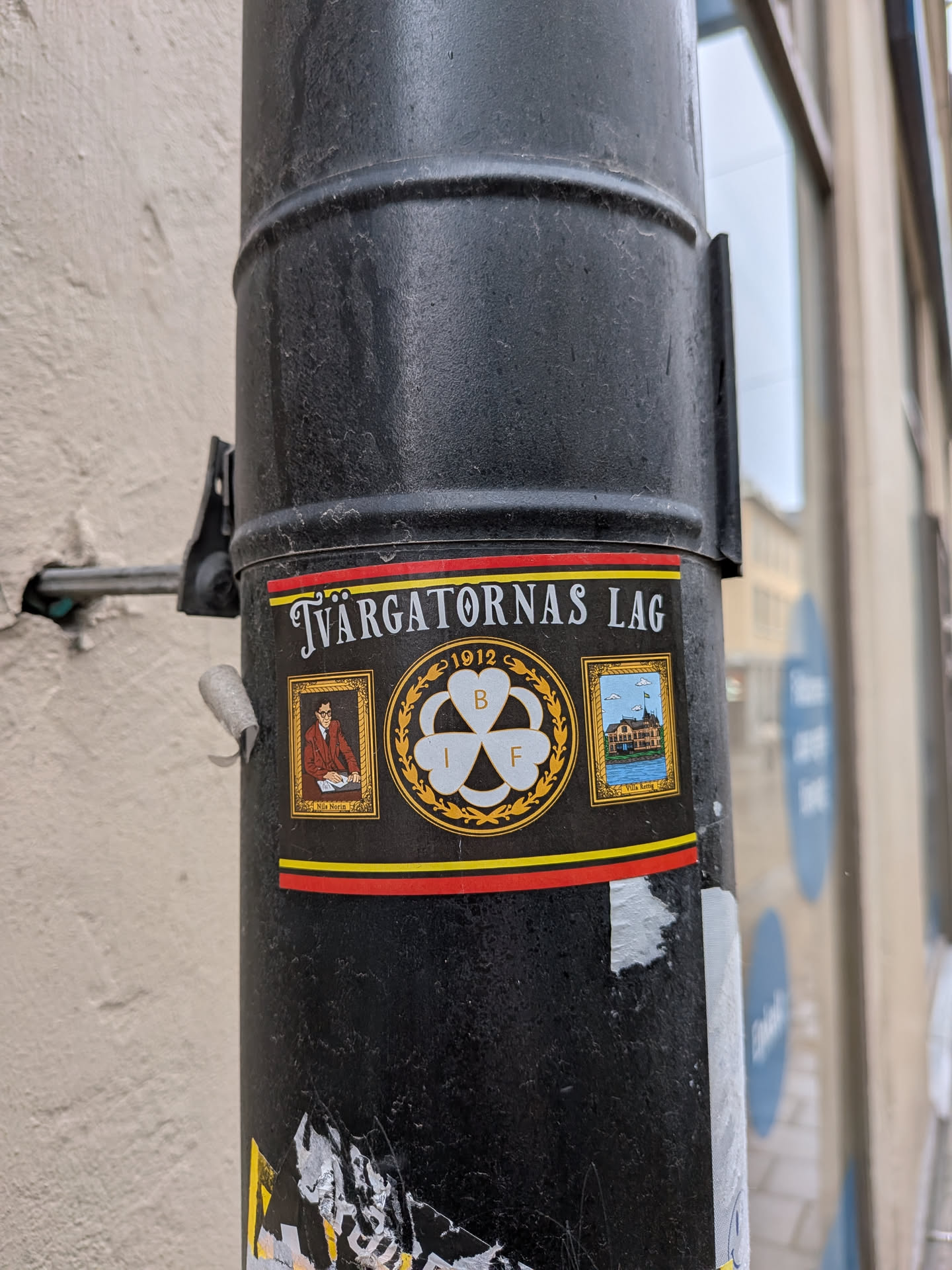 The sticker is rectangular with a black background, bordered by a red stripe at the top and a red and yellow stripe at the bottom. The top features the text 'Tvärgatornas Lag' in white capital letters. Below this, the sticker is divided into three main visual elements. On the left, a framed portrait depicts a man with glasses, wearing a brown jacket, seated at a desk with papers, identified by the name 'Nils Norin' below. In the center, a prominent gold-bordered circular emblem showcases a white three-leaf clover or shamrock, with the letters 'B', 'I', and 'F' inscribed within its leaves, and the year '1912' above. On the right, another framed image displays a painting or illustration of a large, multi-story house or villa situated by water, with a flag flying from its roof, identified as 'Villa Rettig' below. The overall aesthetic is classic and commemorative, utilizing gold and white accents against the dark background.