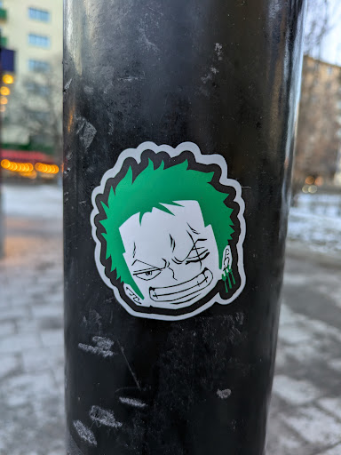 Street sticker A sticker of Zoro from One Piece is stuck on a metal pole. He has green hair, is missing his left eye, and is wearing green earrings.