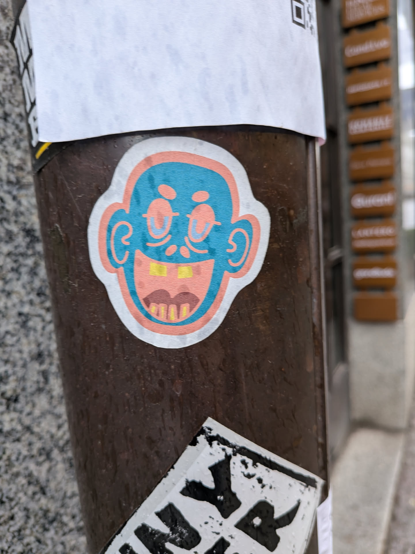 A die-cut sticker featuring a cartoonish head with a wide, open-mouthed grin. The face is predominantly a bright teal blue, outlined in a lighter peach or salmon pink. It has large, round ears, also outlined in peach. The eyes are simple blue ovals with lighter blue pupils, giving a slightly squinted or happy expression. The mouth is wide open, revealing a peach-colored interior, a dark brown upper lip or mustache, and yellow teeth, some of which appear to be missing, creating a gap-toothed look. The entire design is bordered by a thin white outline, suggesting it's a cut-out sticker.