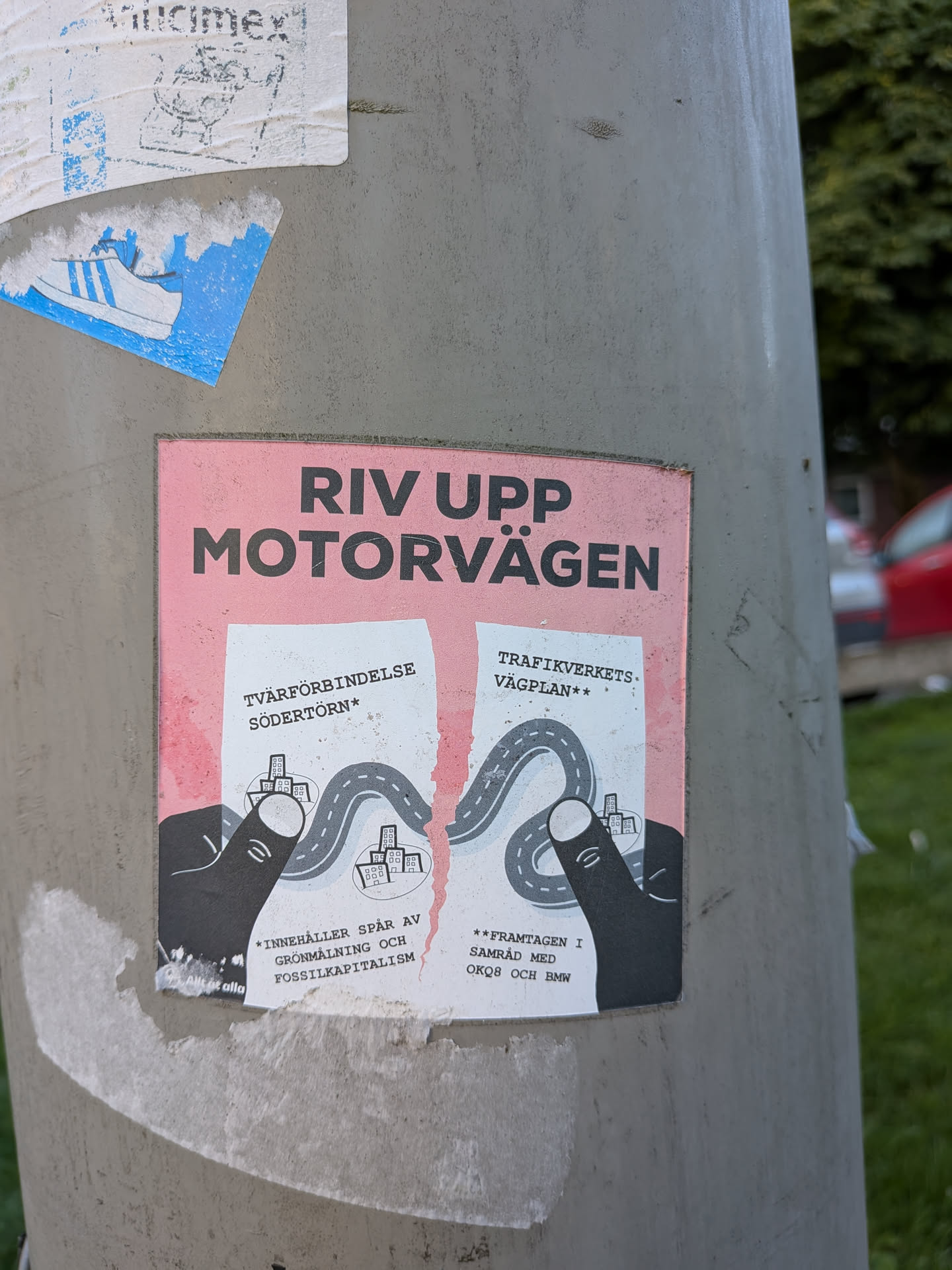 Street sticker The sticker features a pink background with the text "RIV UPP MOTORVÄGEN" in large, bold black letters at the top. Below this, two torn pieces of paper are depicted, each with a road winding through miniature cityscapes. The left piece has text that reads "TVÄRFÖRBINDELSE SÖDERTÖRN*" and below that "*INNEHÅLLER SPÅR AV GRÖNMÅLNING OCH FOSSILKAPITALISM". The right piece reads "TRAFIKVERKETS VÄGPLAN**" and below that "**FRAMTAGEN I SAMRÅD MED OKQ8 OCH BMW". Two black, stylized hands are shown holding and tearing the two pieces of paper apart, illustrating the message of dismantling the highway. The overall aesthetic is somewhat rough and distressed, with subtle speckling on the pink background.