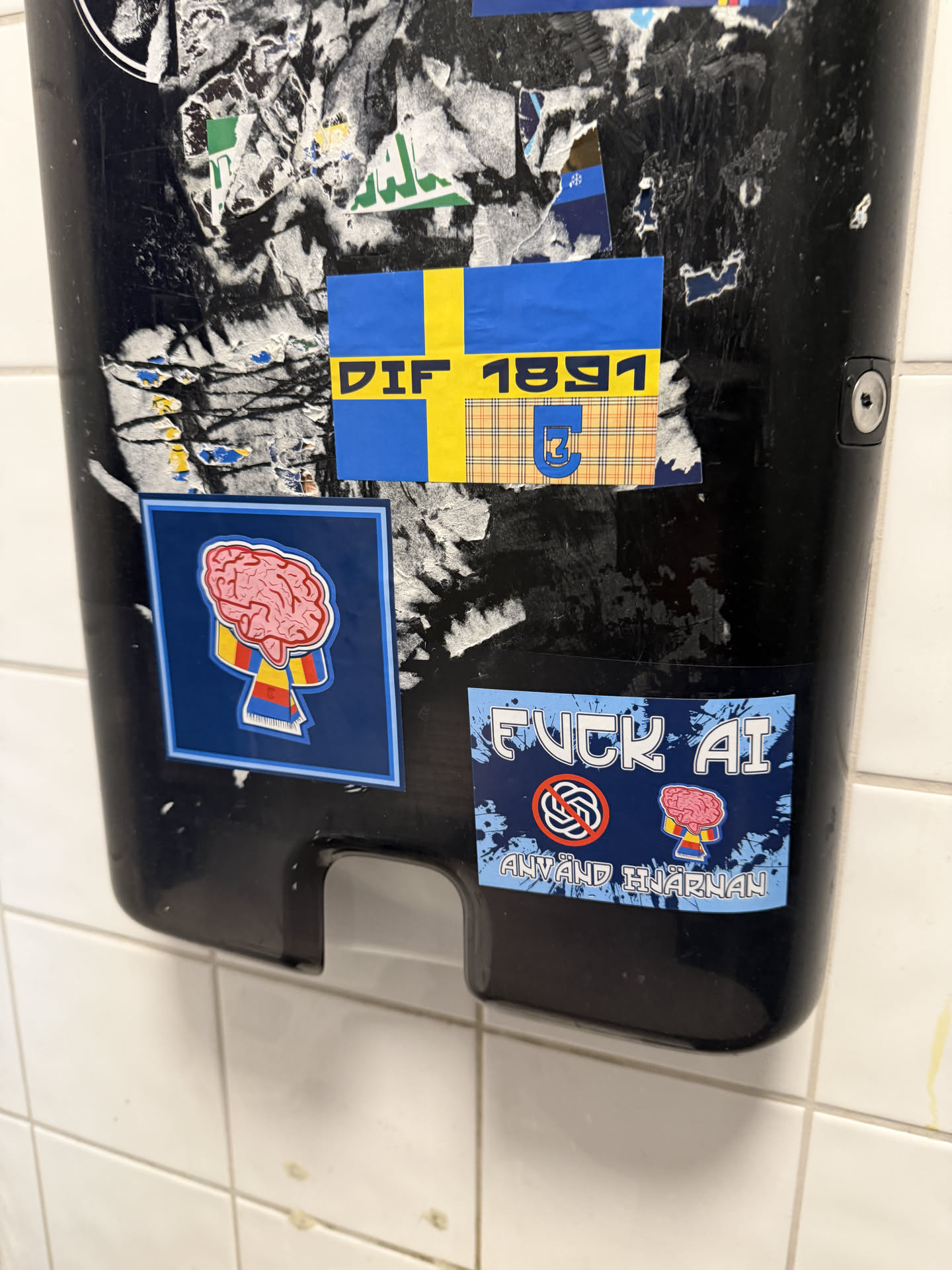 This rectangular sticker features a design inspired by the Swedish flag, with a prominent yellow Nordic cross on a blue background. The top left and bottom left quadrants are solid blue. The top right quadrant is blue with the yellow text 'DIF' above '1891'. The bottom right quadrant displays a brown, yellow, and blue plaid pattern, overlaid with a stylized blue crest or monogram, likely representing the club's emblem. The sticker shows signs of wear and slight peeling, indicating it is genuinely affixed to the surface.