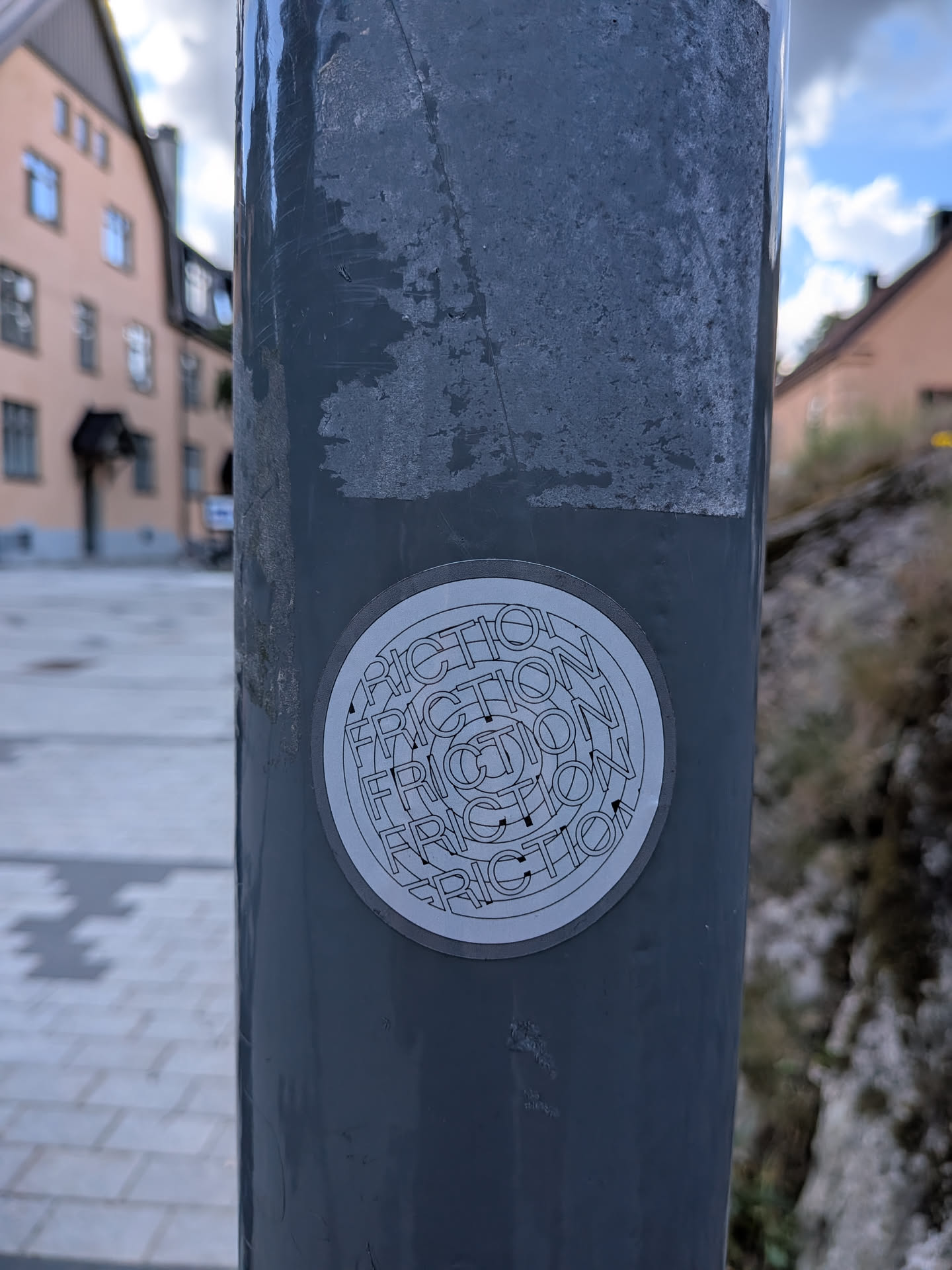 A round grey sticker featuring the word 'FRICTION' repeated in concentric, warped outline text, creating a circular optical pattern.