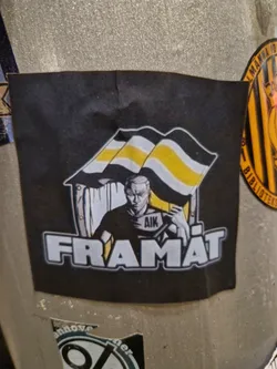 Street sticker A black and yellow sticker with an illustration of a person holding a flag that says AIK. The text "FRAMÅT" is written underneath the illustration.  The style is reminiscent of political or activist art.