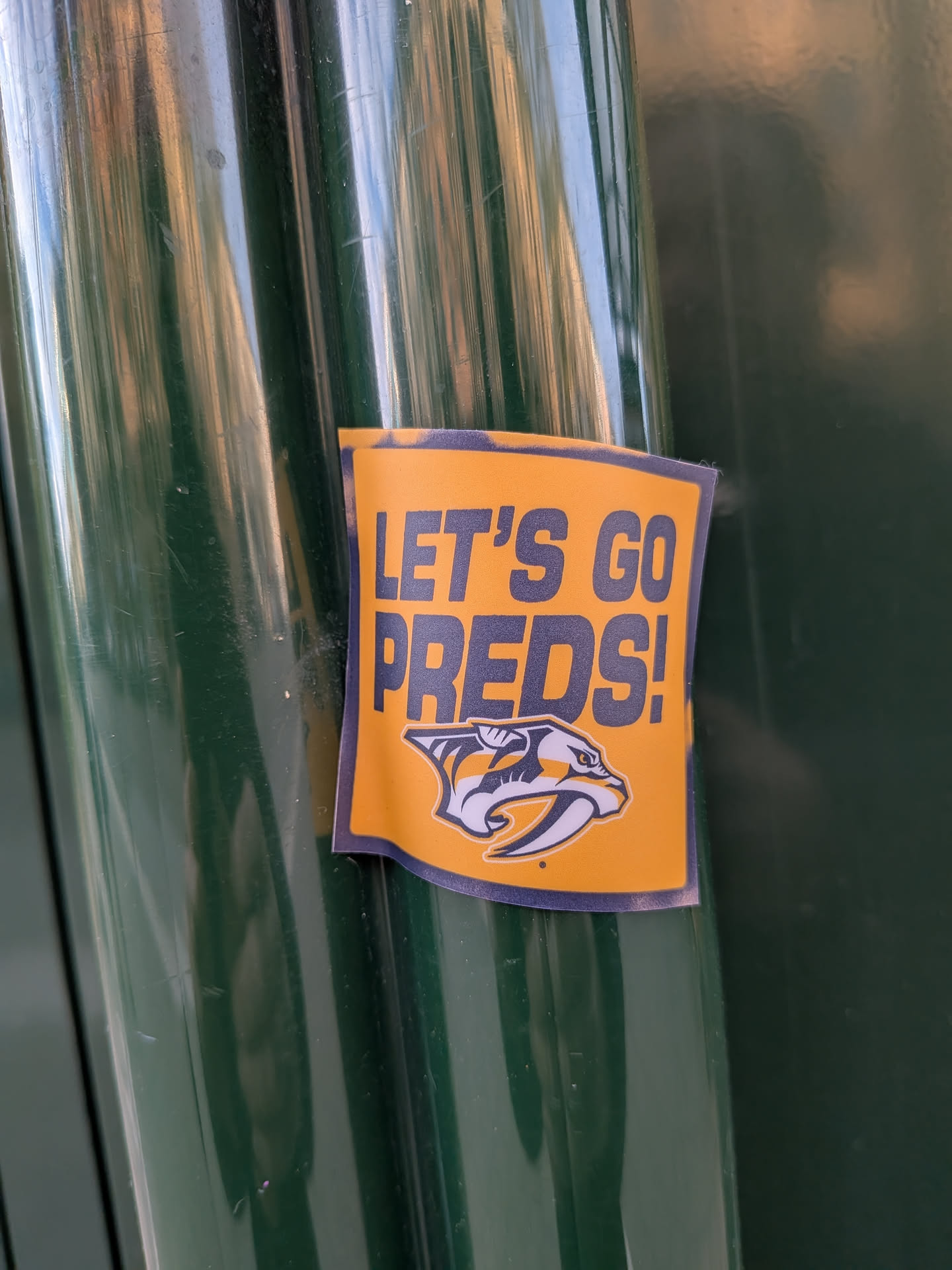Street sticker The sticker is predominantly orange and blue, featuring the text 'LET'S GO PREDS!' in bold, dark blue capital letters. Below the text is the logo of the Nashville Predators hockey team, which depicts a stylized saber-toothed cat's head in white and yellow with blue accents. The sticker has a slightly glossy finish and appears to be adhered to a dark green, cylindrical surface.