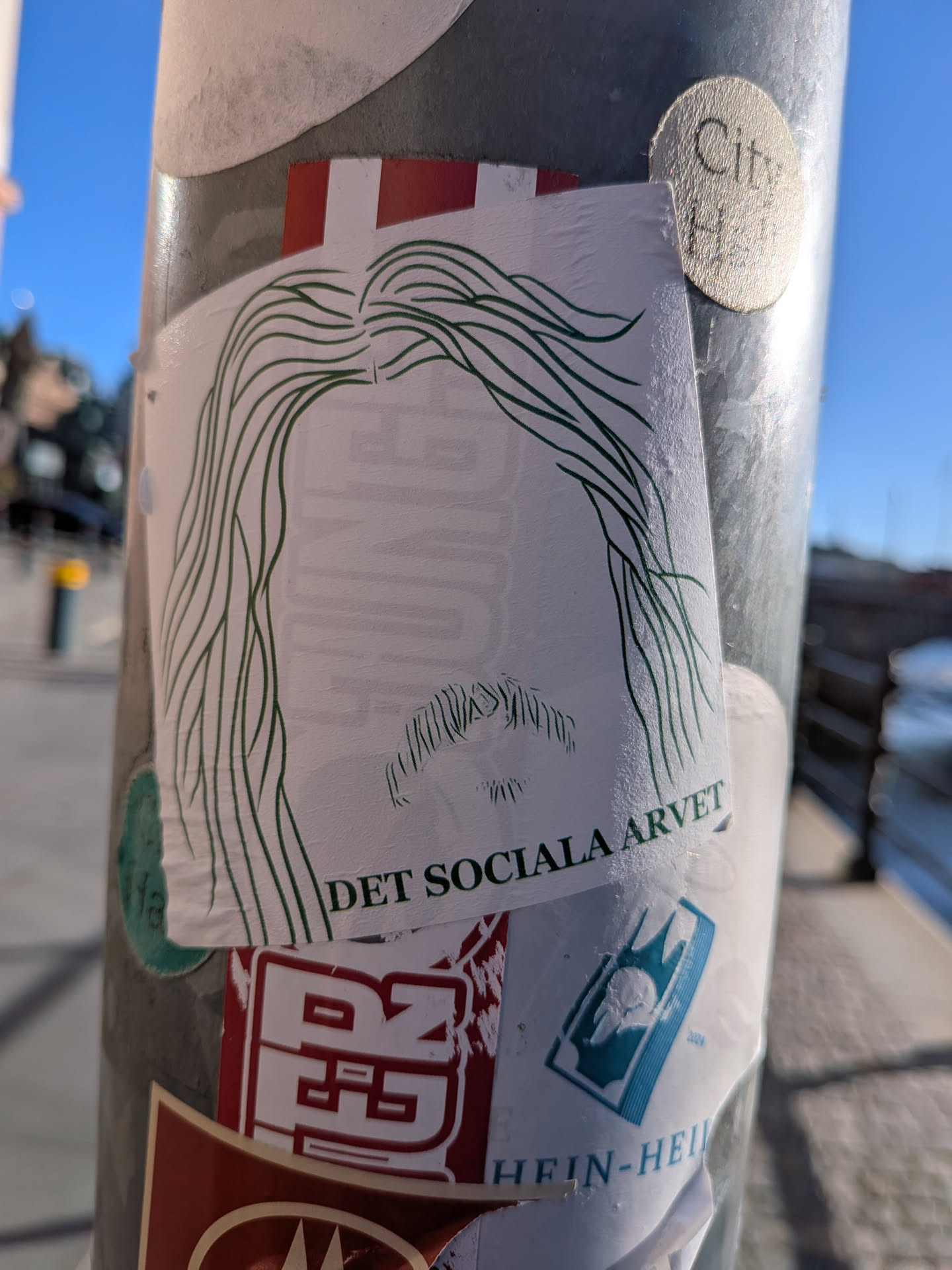 The sticker is predominantly white with a green line drawing of a person's head and shoulders. The hair is depicted as long, flowing strands of green lines. A mustache is visible below the nose area. The text 'DET SOCIALA ARVET' is printed in dark green capital letters below the drawing. There are also faint, partially obscured elements of other stickers around it, including a red and white striped section at the top and a teal graphic with a crown-like symbol and the year '2024' to the right.