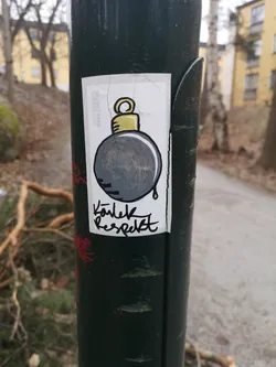 Street sticker A rectangular white sticker adhered to a dark green pole. The central image is a stylized, cartoonish bomb with a thick black outline. The bomb's body is filled with a dark grey, almost scribbled texture, and a small black drip extends from its bottom. At the top, a yellow, metallic-looking fuse or cap is depicted, also outlined in black. Below the bomb, in a flowing black handwritten script, are two words. Along the left edge of the sticker, oriented vertically, a faint logo or brand name is visible. The sticker itself shows signs of wear, with several fine cracks across its surface.