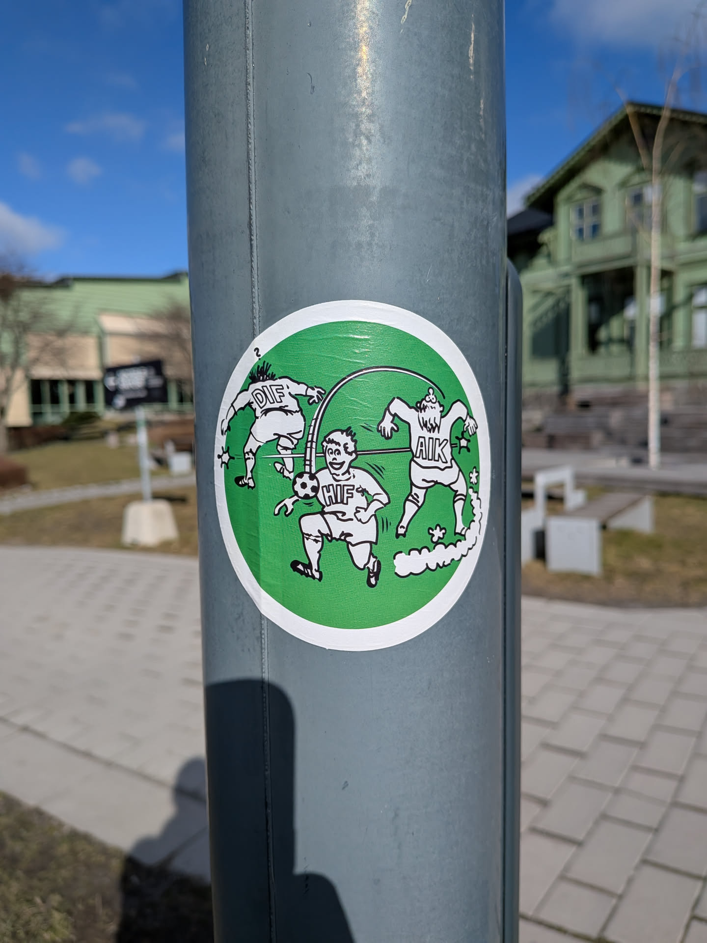 A circular sticker with a vibrant green background and a clean white border. It features three stylized, cartoonish male figures in white outfits with black outlines, engaged in a dynamic football scene. The figure on the left, with spiky hair, is shown mid-kick with 'DIF' written on his back. The central figure, smiling, holds a football and has 'HIF' on his chest. The figure on the right, with a beard and spiky hair, is in motion, with 'AIK' on his back, leaving a trail of white smoke or dust. A thin black line connects the figures, suggesting continuous action. Small white stars and other decorative elements are scattered around the characters, adding to the energetic and humorous feel.