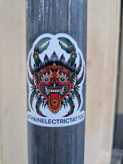 Street sticker A sticker featuring a traditional tattoo-style illustration of a crab with a demonic face in the center. The crab is depicted with green claws and a red, detailed face. Below the illustration, the text "@chainelectrictattoo" is visible.