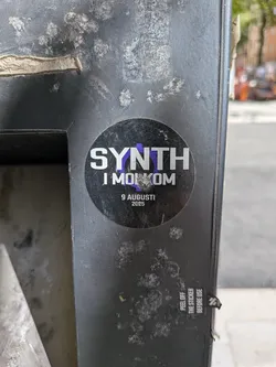 Street sticker A circular sticker with a dark, possibly black or very dark grey, background. The main text 'SYNTH' is prominently displayed in large, bold, white sans-serif letters. The 'N' in 'SYNTH' is partially overlaid with a stylized purple graphic, resembling a gear or an abstract symbol. Below this, in smaller white sans-serif font, is 'I MOL KOM'. Further below, centered, the date '9 AUGUSTI' and '2025' are written in even smaller white sans-serif text. The sticker shows some minor scuffs and wear on its surface. A 'PEEL OFF THE STICKER BEFORE USE' tab is visible at the bottom right, indicating it's a new sticker yet to be fully applied or removed from its backing.