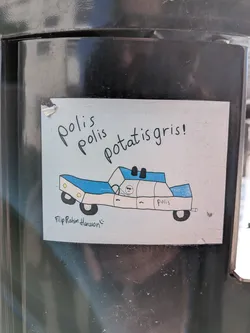 Street sticker The sticker is rectangular with a light blue background. At the top, in black handwritten text, it reads 'polis polis potatis gris!'. Below the text is a hand-drawn image of a police car. The car is predominantly white with blue accents on the roof and sides. It has black wheels and yellow circular details on the front. A small, stick-figure-like character with sunglasses is drawn inside the car. The word 'polis' is written on the side of the car. Below the car, in smaller cursive handwriting, is 'Filip Rahim Hansson' followed by a smiley face emoji. The sticker appears to be slightly worn at the edges.