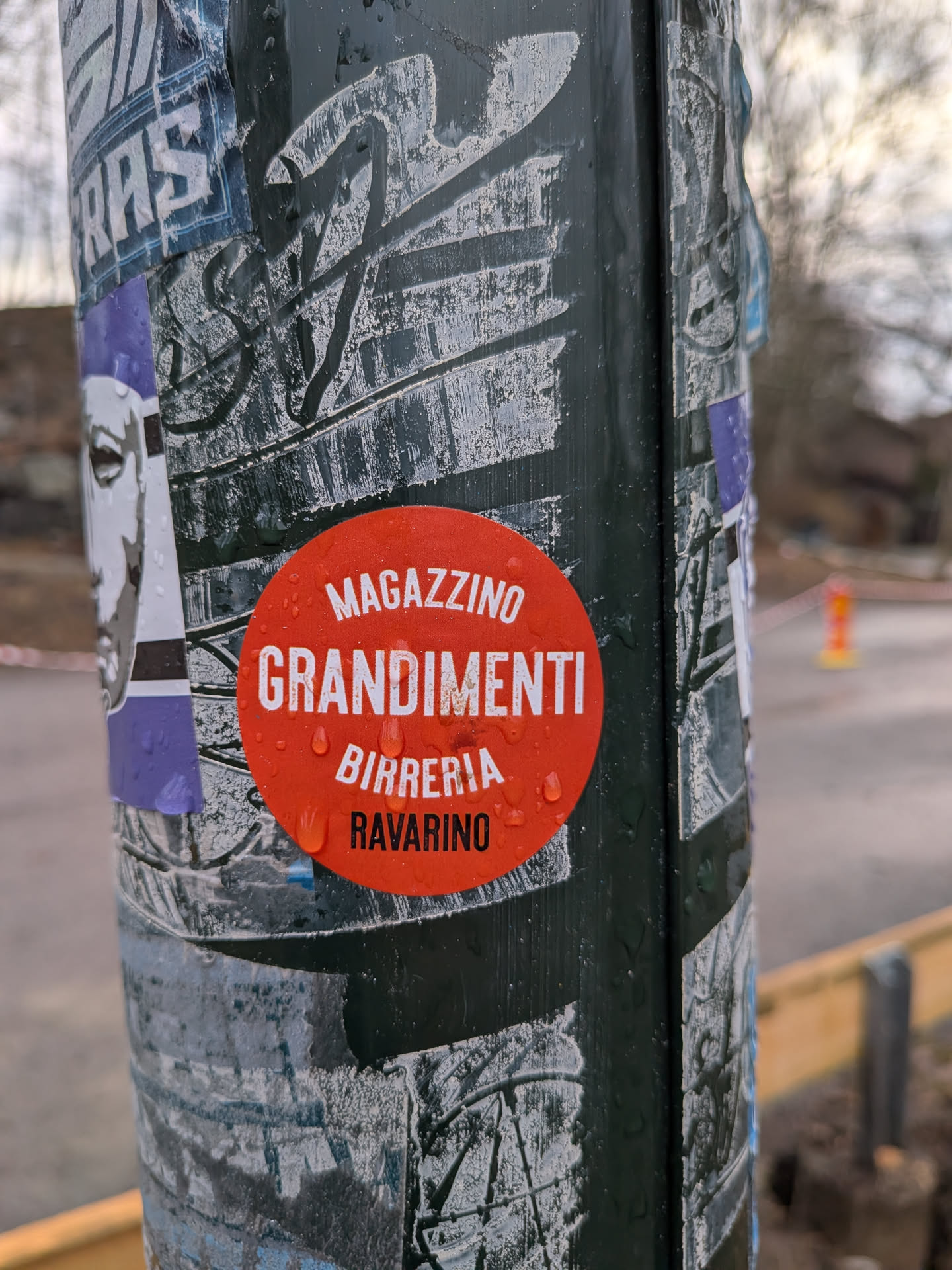 The sticker is circular and predominantly red, with a matte finish. It features white sans-serif text arranged in three main lines: 'MAGAZZINO' at the top, 'GRANDIMENTI' as the largest and central text, and 'BIRRERIA' below it. A fourth, smaller line of text in black, 'RAVARINO', is positioned at the very bottom. The sticker is wet, with numerous small water droplets visible on its surface, suggesting it is outdoors and exposed to the elements. It is affixed to a dark, possibly green or black, pole that is also covered with other faded and partially torn stickers and graffiti.