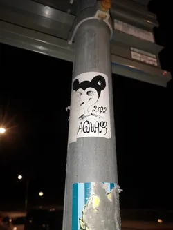 Street sticker The sticker is a roughly rectangular piece of white paper, slightly peeling at the edges, affixed to a metal pole. Its central design is a black and white illustration of a stylized head. At the top, there are two prominent, solid black, rounded ears, strongly reminiscent of Mickey Mouse. Below the ears, the face is depicted with a more human-like, somewhat elongated form. The most striking feature of the face is a large, wide-open eye with a distinct pupil and iris, drawn with fine black lines on the white background. The nose and mouth are not clearly defined, giving the face an abstract or minimalist quality. To the right of the face, the year '2022' is written in a casual, slightly cursive script. Below the year, the name or tag 'AGNAR' is written in a bold, stylized, almost graffiti-like font, with some letters incorporating musical note-like elements. The overall aesthetic is that of a hand-drawn, urban art piece.
