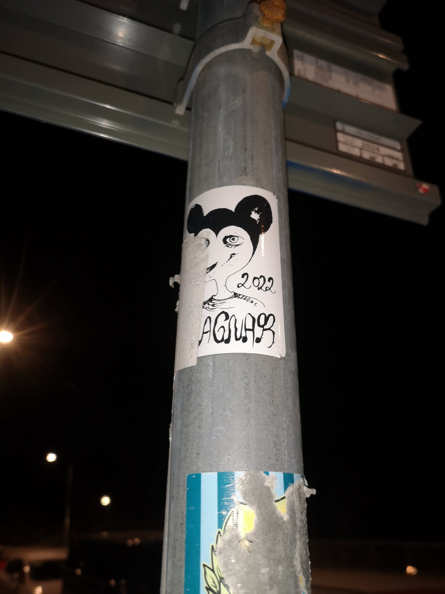 The sticker is a roughly rectangular piece of white paper, slightly peeling at the edges, affixed to a metal pole. Its central design is a black and white illustration of a stylized head. At the top, there are two prominent, solid black, rounded ears, strongly reminiscent of Mickey Mouse. Below the ears, the face is depicted with a more human-like, somewhat elongated form. The most striking feature of the face is a large, wide-open eye with a distinct pupil and iris, drawn with fine black lines on the white background. The nose and mouth are not clearly defined, giving the face an abstract or minimalist quality. To the right of the face, the year '2022' is written in a casual, slightly cursive script. Below the year, the name or tag 'AGNAR' is written in a bold, stylized, almost graffiti-like font, with some letters incorporating musical note-like elements. The overall aesthetic is that of a hand-drawn, urban art piece.