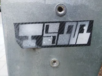 A black and white sticker with an abstract design, possibly representing buildings or structures. It's applied to a metallic surface. The sticker has a carbon fiber-like texture.