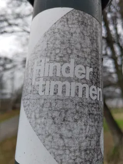 A worn-out sticker with the word "Hindertimmen" printed on it. The sticker is affixed to a dark-colored pole outdoors, showing significant signs of weathering and dirt.